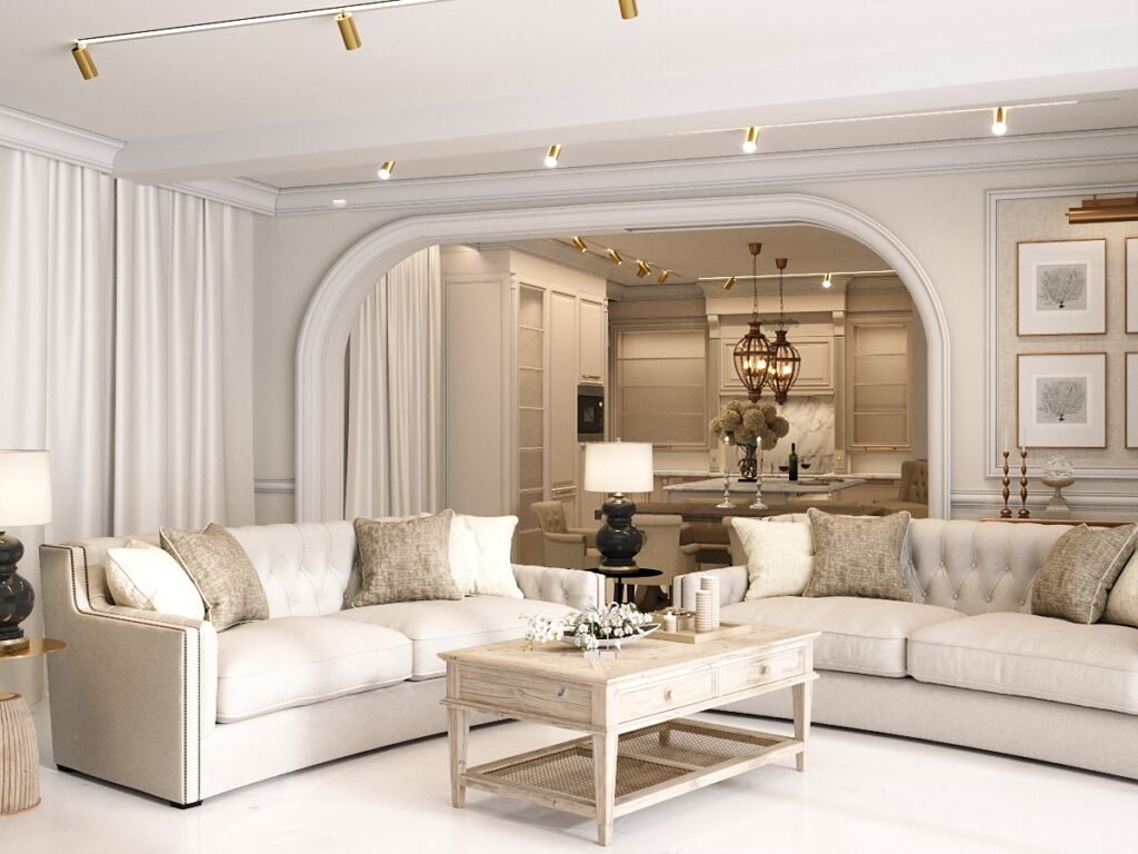 Elegancki salon w jasnych kolorach z dużą narożną sofą w beżowych odcieniach, drewnianym stolikiem kawowym i poduszkami w naturalnych tonach. W tle widoczna otwarta kuchnia z wyspą, a całość oświetlona nowoczesnym oświetleniem sufitowym.